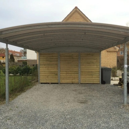 Carport med skur i træ og buet tagkonstruktion på grusbelægning.